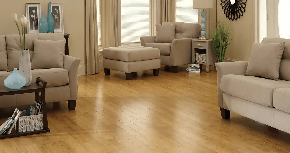 A living room with professionally installed bamboo flooring, beige sofas, a matching ottoman, a side table with a blue lamp, vases, framed pictures, and large windows with blinds and curtains in Stockton, CA